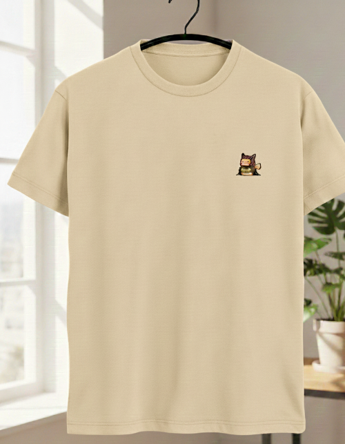 Beige t-shirt with a small graphic on a hanger in a room with a plant.