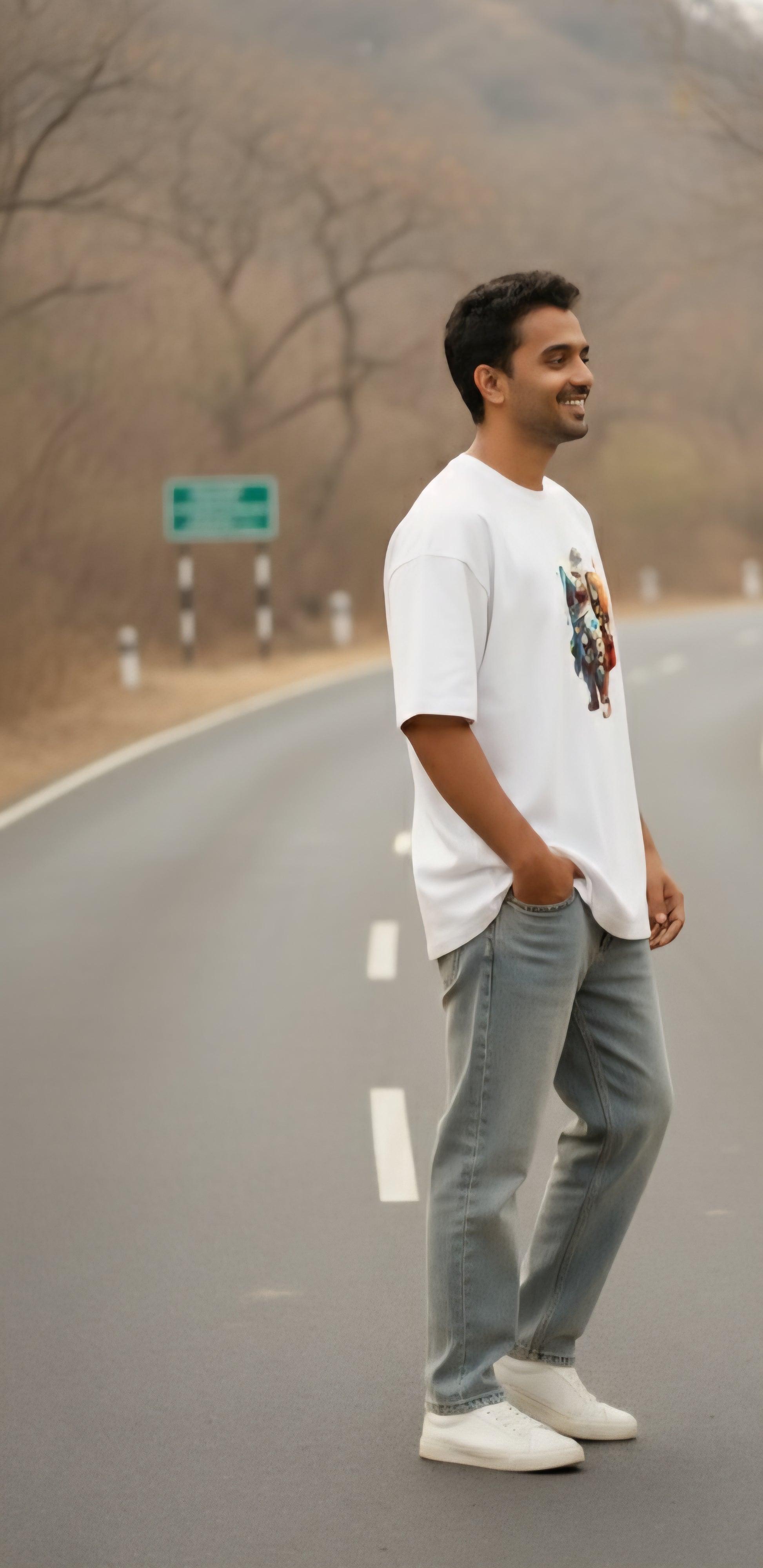 Man in white graphic t-shirt and jeans standing on empty road with trees in background