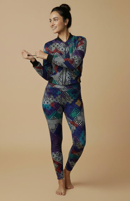 Women's printed activewear co-ord set featuring a colorful geometric bomber jacket and matching leggings, worn by a smiling female model standing barefoot against a beige studio background — iTiDharma athleisure fashion.