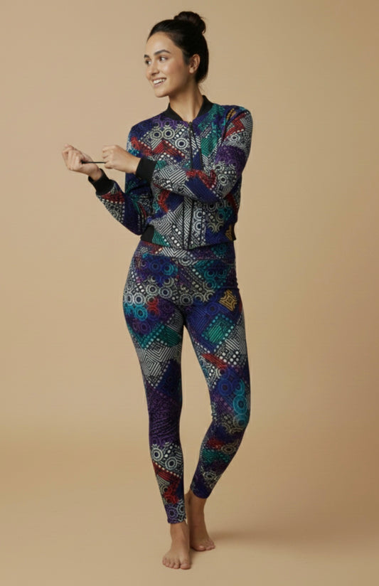 Women's printed activewear co-ord set featuring a colorful geometric bomber jacket and matching leggings, worn by a smiling female model standing barefoot against a beige studio background — iTiDharma athleisure fashion.